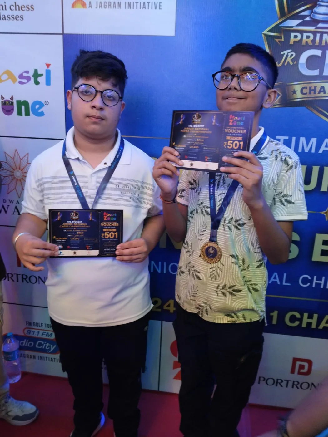 Two players with their medals and Masti Zone vouchers in front of the Prime Engage Jr. Chess Championship partner wall in Delhi