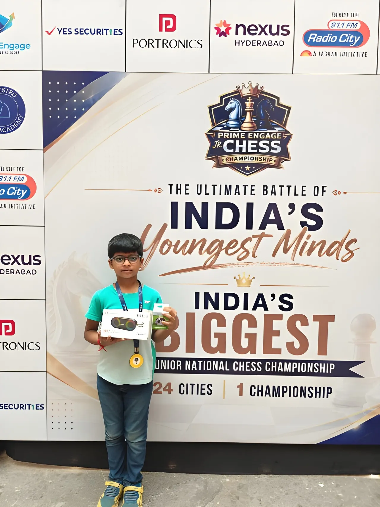 A young player with a medal and a gadget prize in front of the championship backdrop, which carries the Nexus Hyderabad logo.